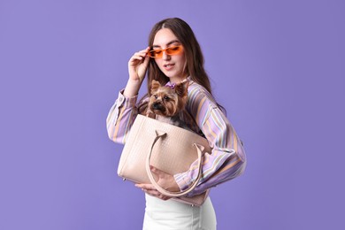 Beautiful young woman carrying cute Yorkshire Terrier dog in bag on violet background