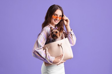 Photo of Beautiful young woman carrying cute Yorkshire Terrier dog in bag on violet background