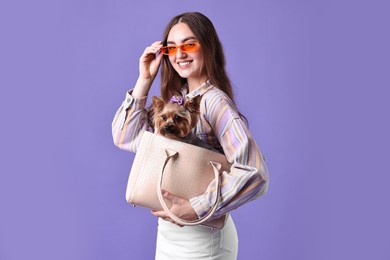 Photo of Beautiful young woman carrying cute Yorkshire Terrier dog in bag on violet background