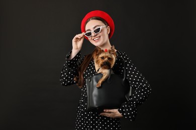 Photo of Woman in sunglasses holding bag with cute Yorkshire Terrier dog on black background