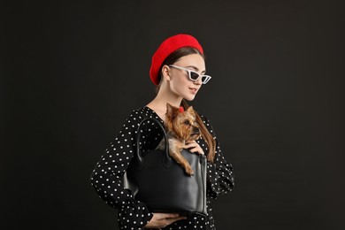 Photo of Woman in sunglasses holding bag with cute Yorkshire Terrier dog on black background