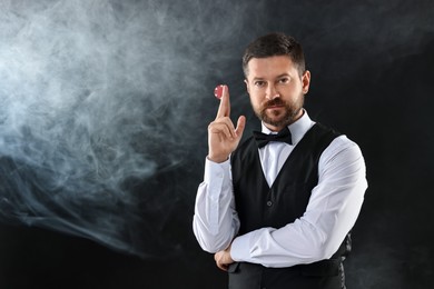 Croupier holding casino chip on black background with smoke, space for text