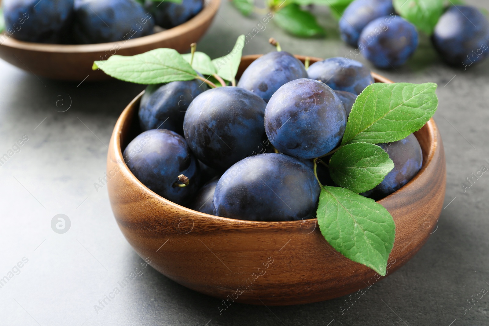 Photo of Tasty ripe plums and leaves in bowl on grey table, closeup