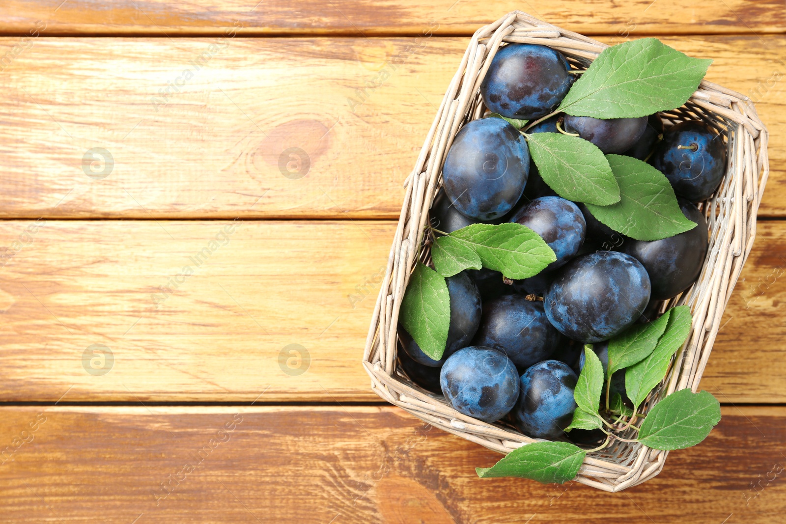 Photo of Tasty ripe plums and leaves in wicker basket on wooden table, top view. Space for text