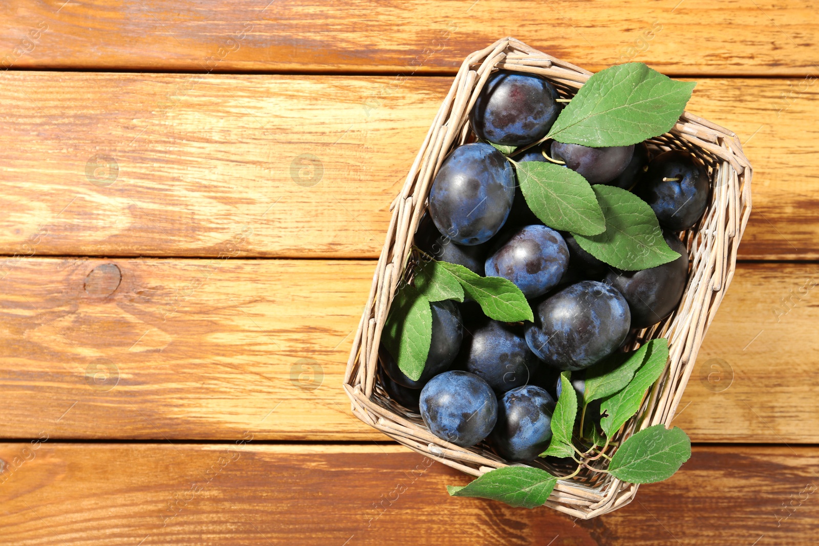 Photo of Tasty ripe plums and leaves in wicker basket on wooden table, top view. Space for text