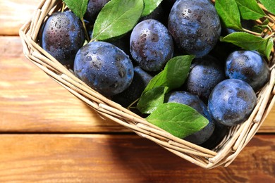 Photo of Tasty ripe plums and leaves in wicker basket on wooden table, top view