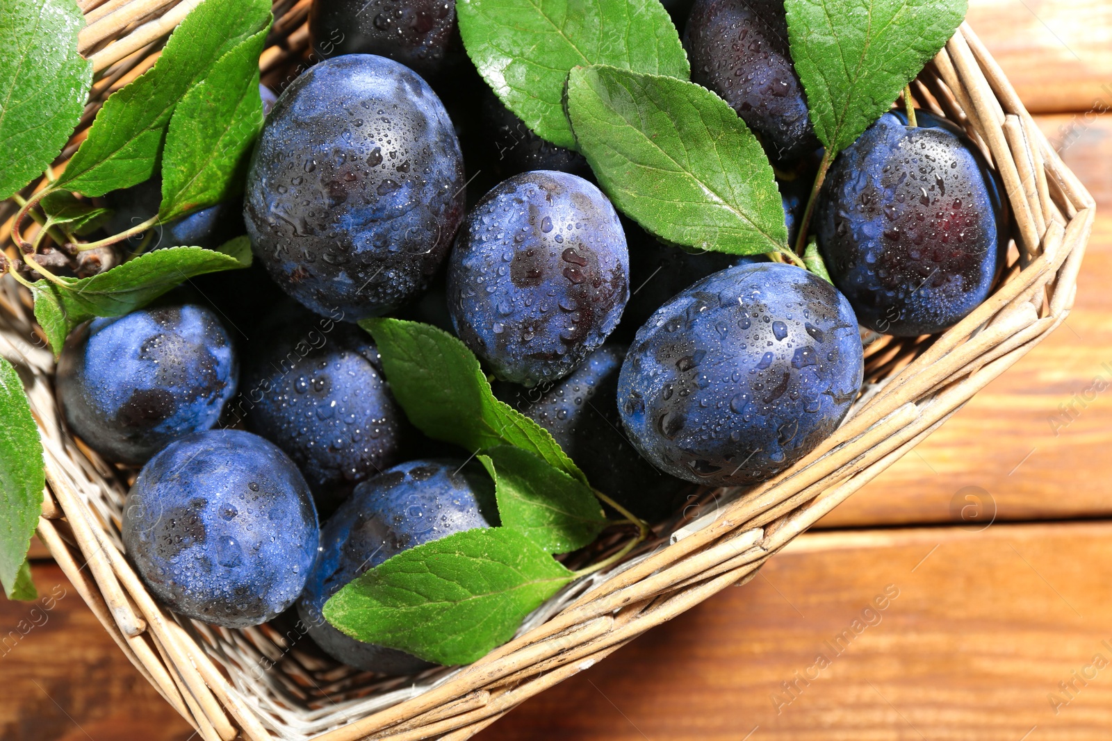 Photo of Tasty ripe plums and leaves in wicker basket on wooden table, top view