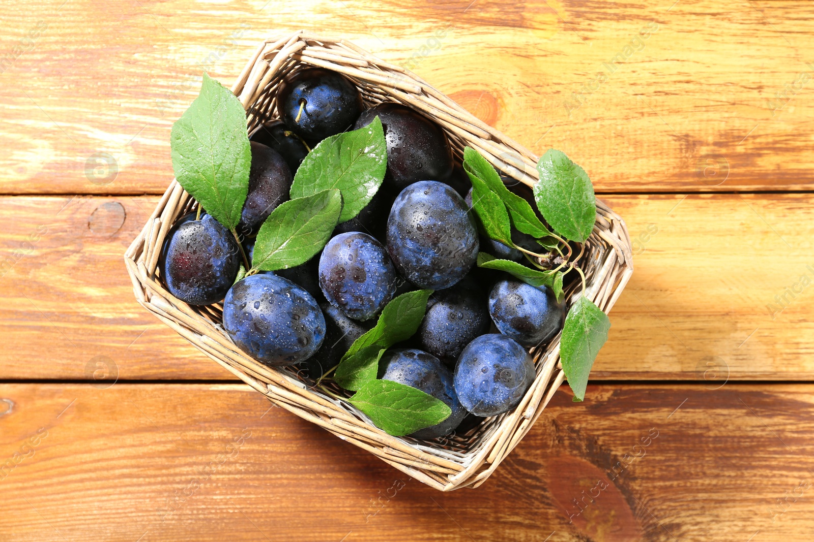 Photo of Tasty ripe plums and leaves in wicker basket on wooden table, top view