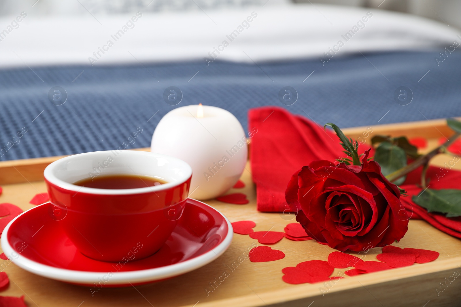 Photo of Wooden tray with burning candle, rose, cup of tea and red paper hearts on bed indoors, closeup