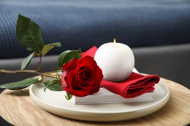 Photo of Burning candle, napkin, red rose and book on wooden table indoors