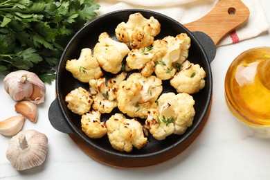 Photo of Tasty baked cauliflower in baking pan, garlic, parsley and oil on white marble table, flat lay