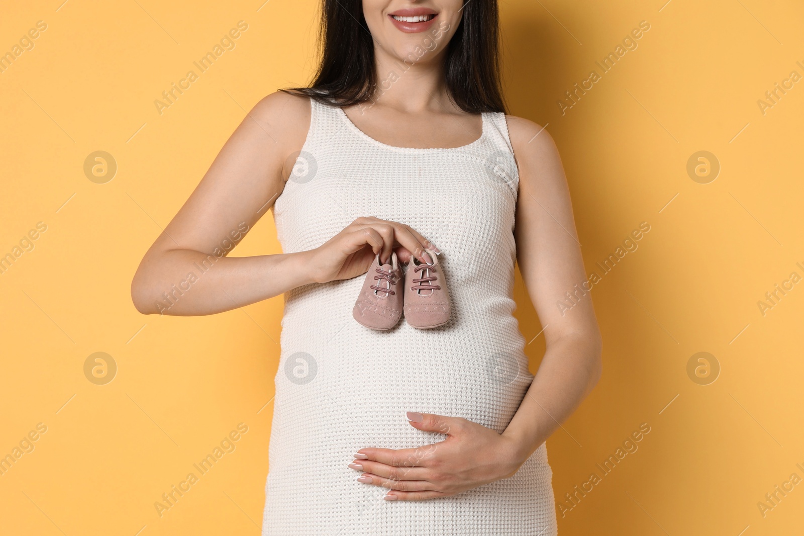 Photo of Young pregnant woman with pair of baby shoes on yellow background, closeup