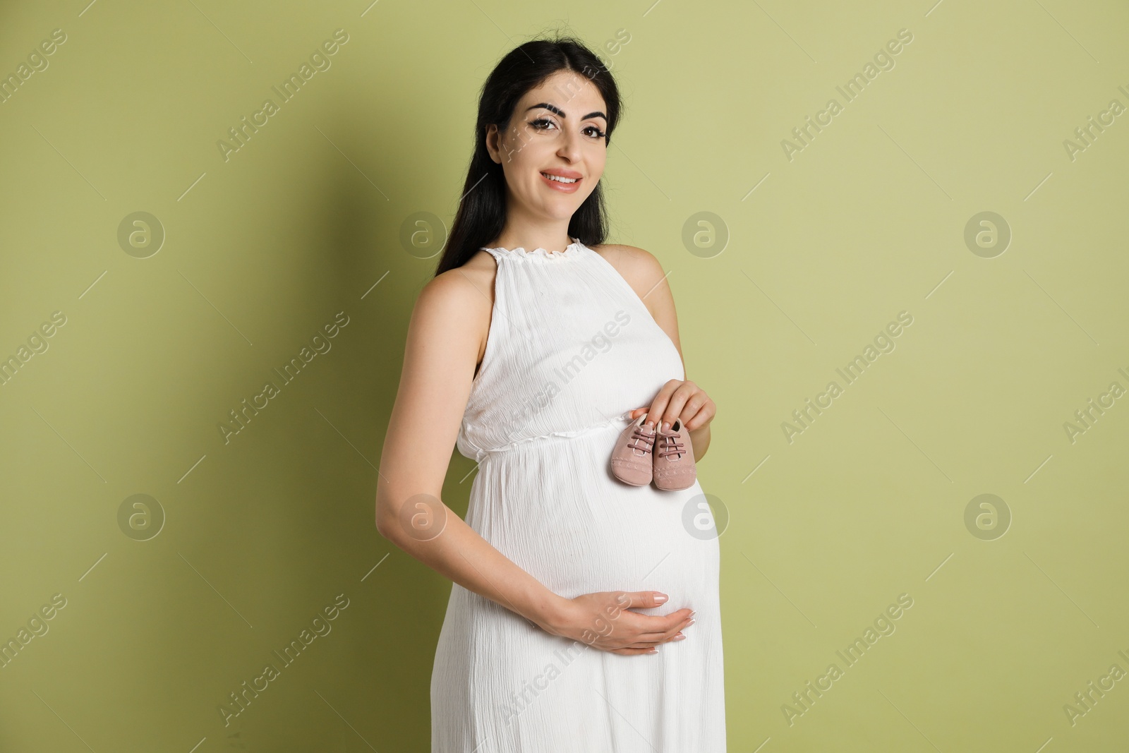 Photo of Portrait of beautiful pregnant woman with baby shoes on light olive background