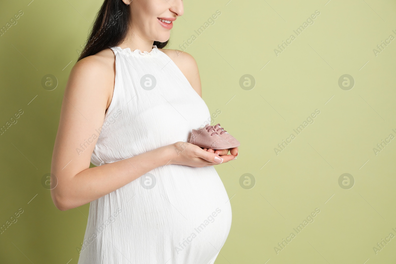 Photo of Young pregnant woman with pair of baby shoes on light olive background, closeup. Space for text