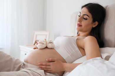 Photo of Young pregnant woman with baby shoes lying on bed at home