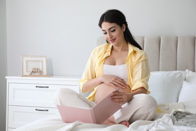 Photo of Young pregnant woman with book sitting on bed at home