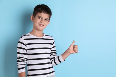 Cute little boy showing thumbs up on light blue background, space for text