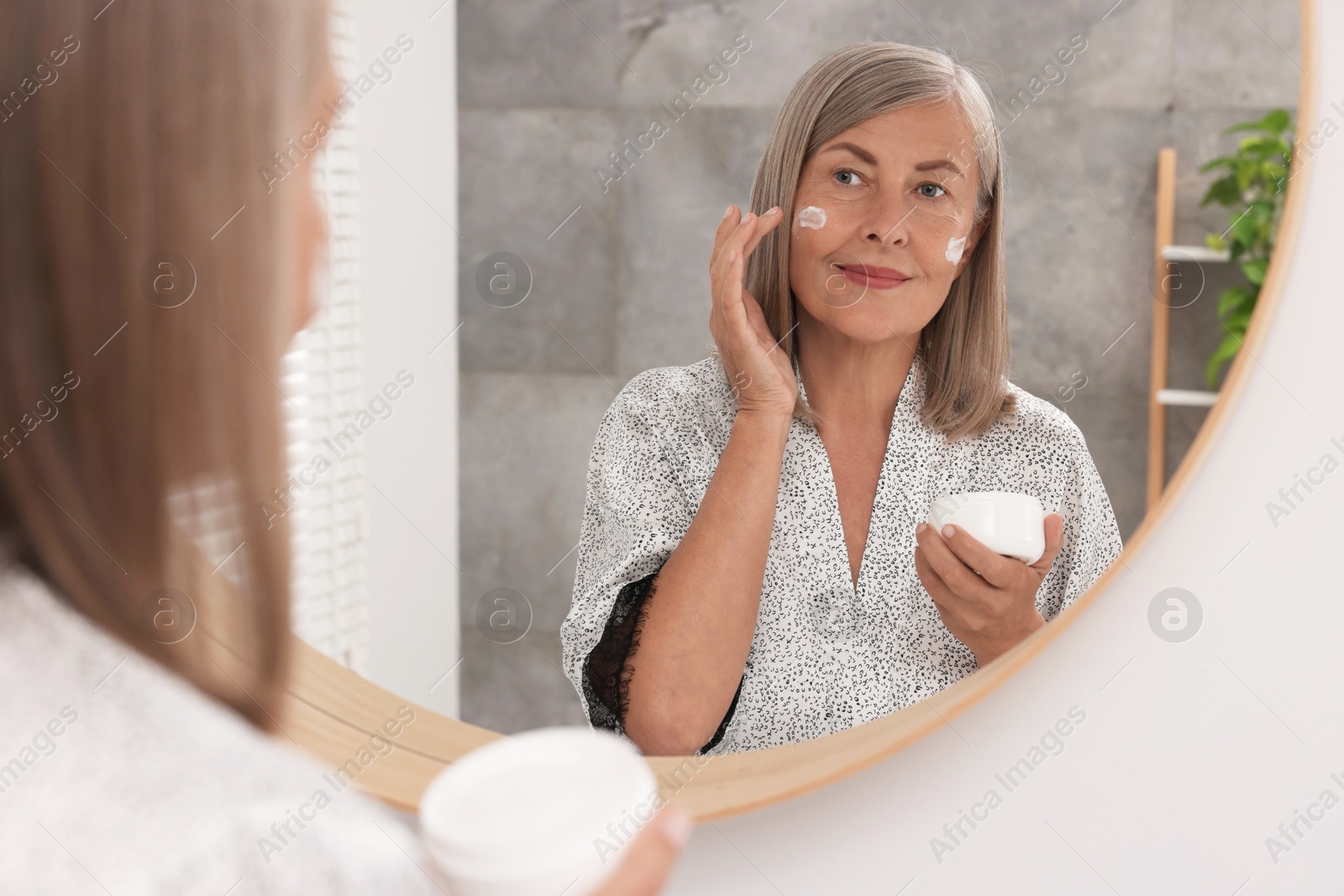 Photo of Senior woman applying face cream near mirror at home