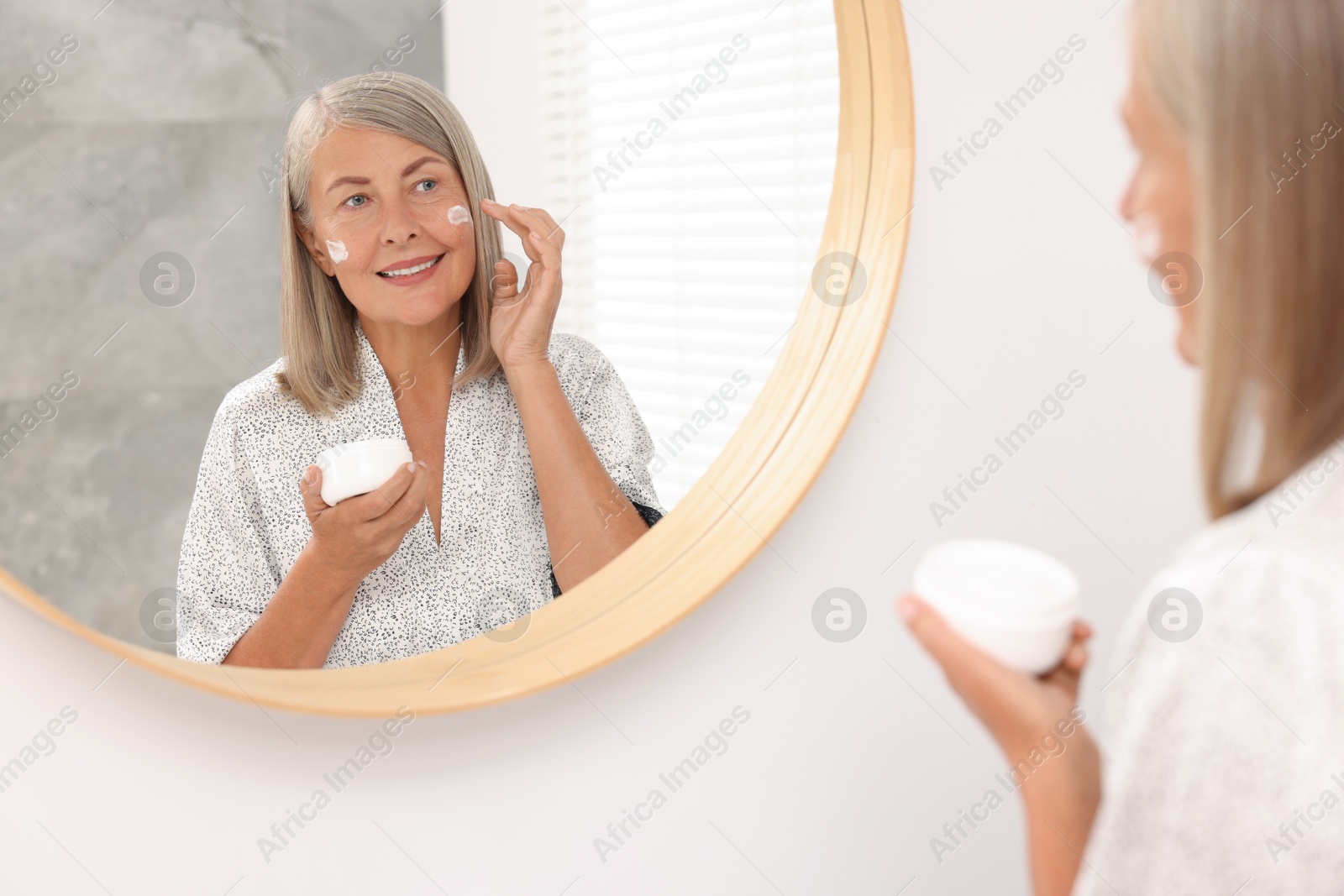 Photo of Senior woman applying face cream near mirror at home