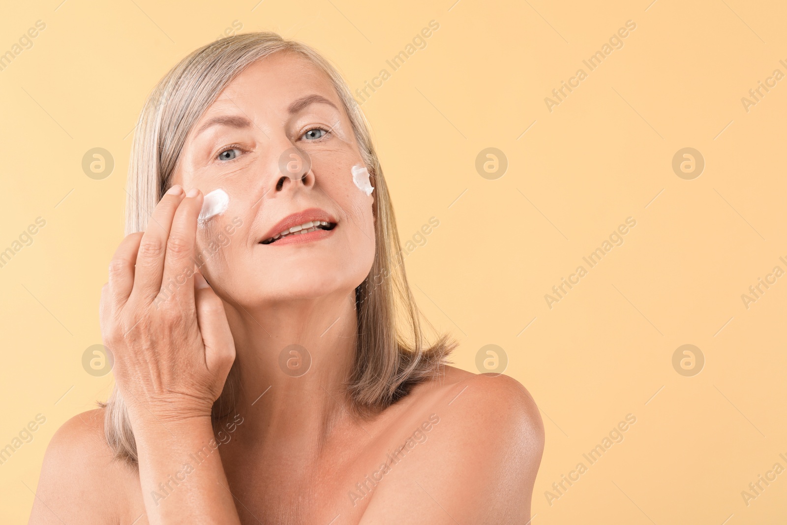 Photo of Senior woman applying face cream on beige background. Space for text