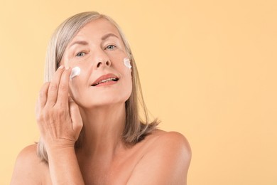 Photo of Senior woman applying face cream on beige background. Space for text