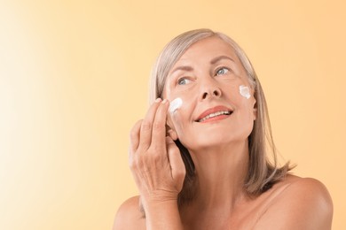 Senior woman applying face cream on beige background. Space for text