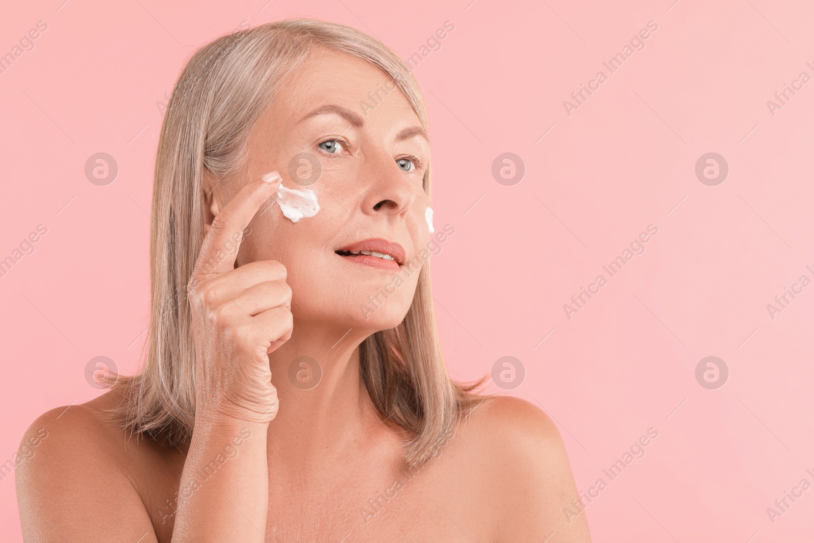 Photo of Senior woman applying face cream on pink background. Space for text