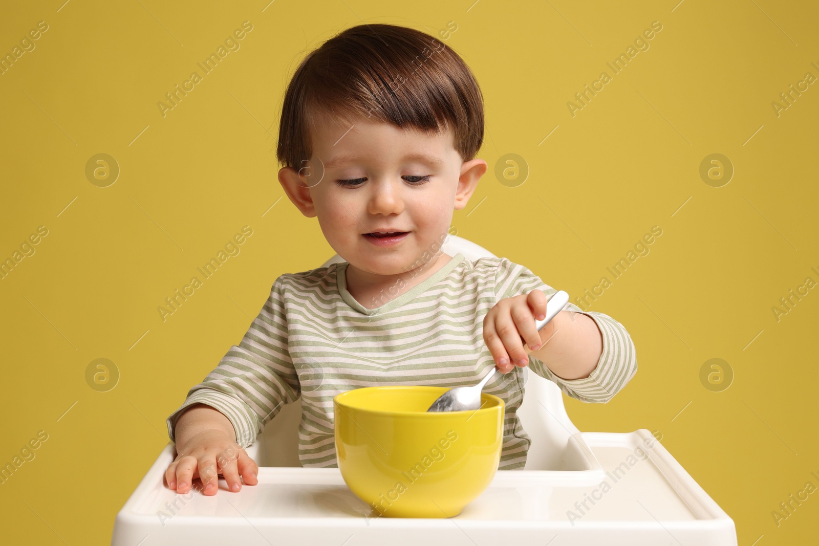 Photo of Cute little kid eating healthy baby food from bowl in high chair on yellow background