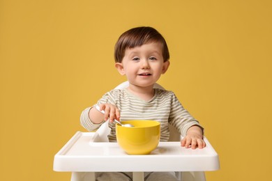 Photo of Cute little kid eating healthy baby food from bowl in high chair on yellow background