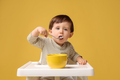 Photo of Cute little kid eating healthy baby food from bowl in high chair on yellow background