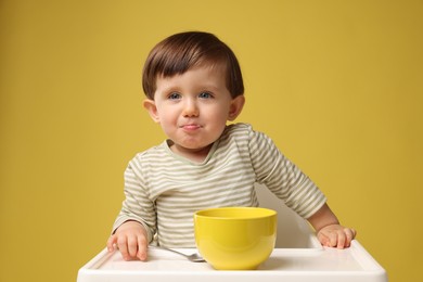 Photo of Cute little kid eating healthy baby food from bowl in high chair on yellow background