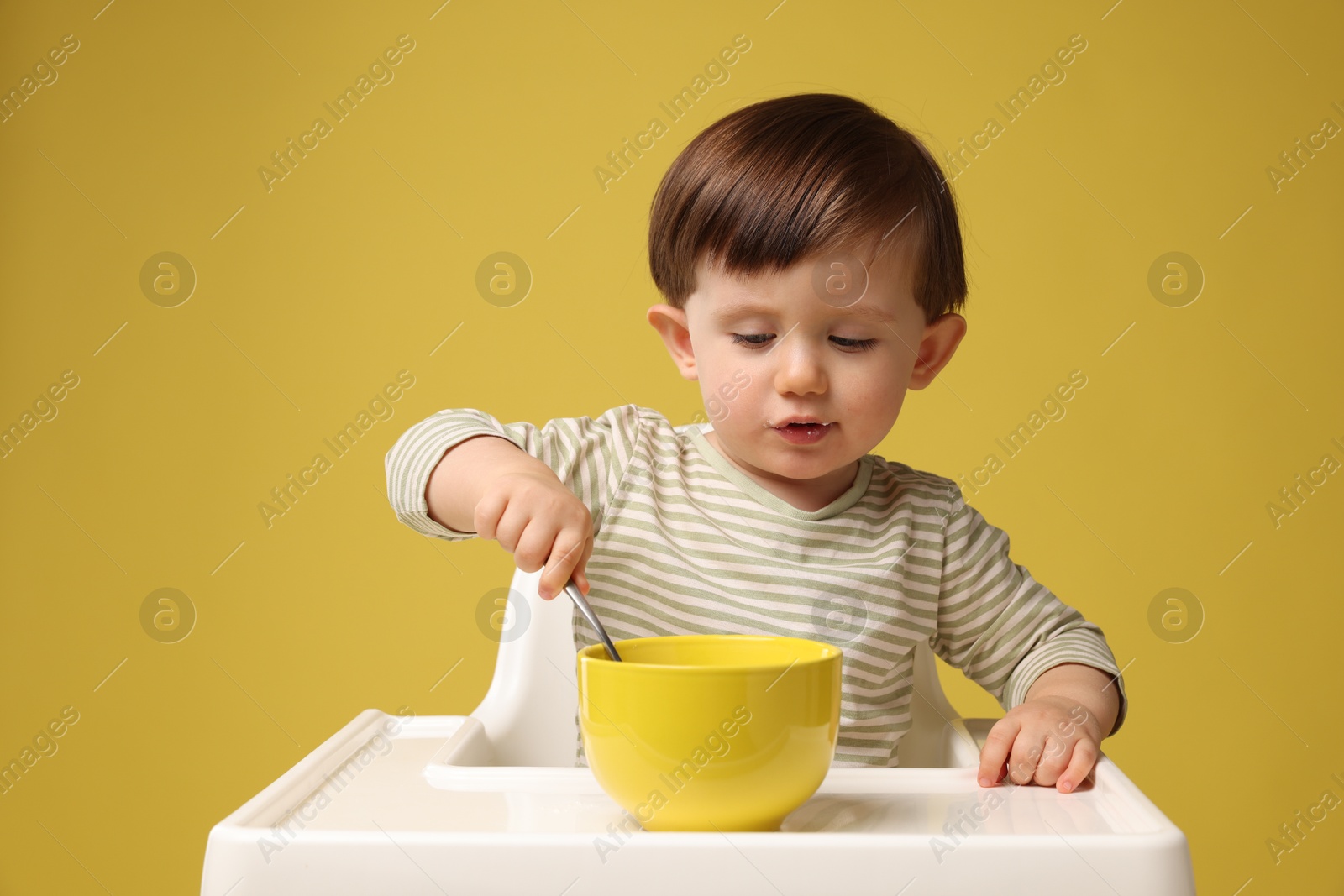 Photo of Cute little kid eating healthy baby food from bowl in high chair on yellow background