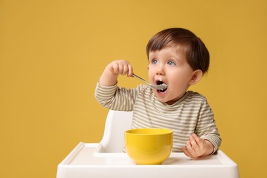 Cute little kid eating healthy baby food from bowl in high chair on yellow background