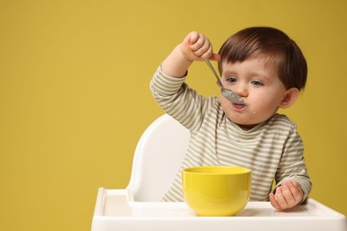 Photo of Cute little kid eating healthy baby food from bowl in high chair on yellow background