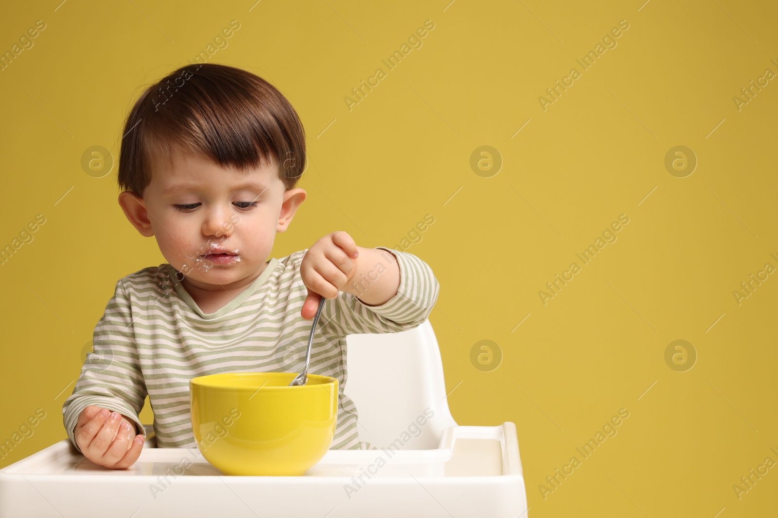 Photo of Cute little kid eating healthy baby food from bowl in high chair on yellow background, space for text