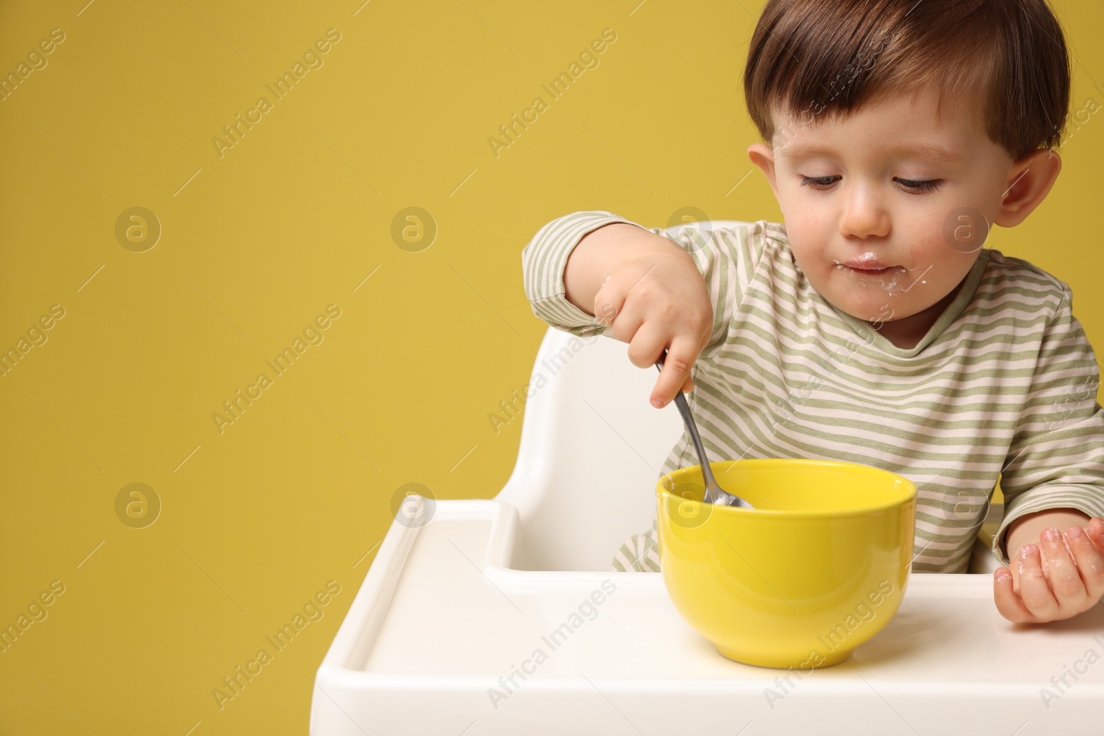 Photo of Cute little kid eating healthy baby food from bowl in high chair on yellow background, space for text