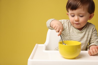 Photo of Cute little kid eating healthy baby food from bowl in high chair on yellow background, space for text