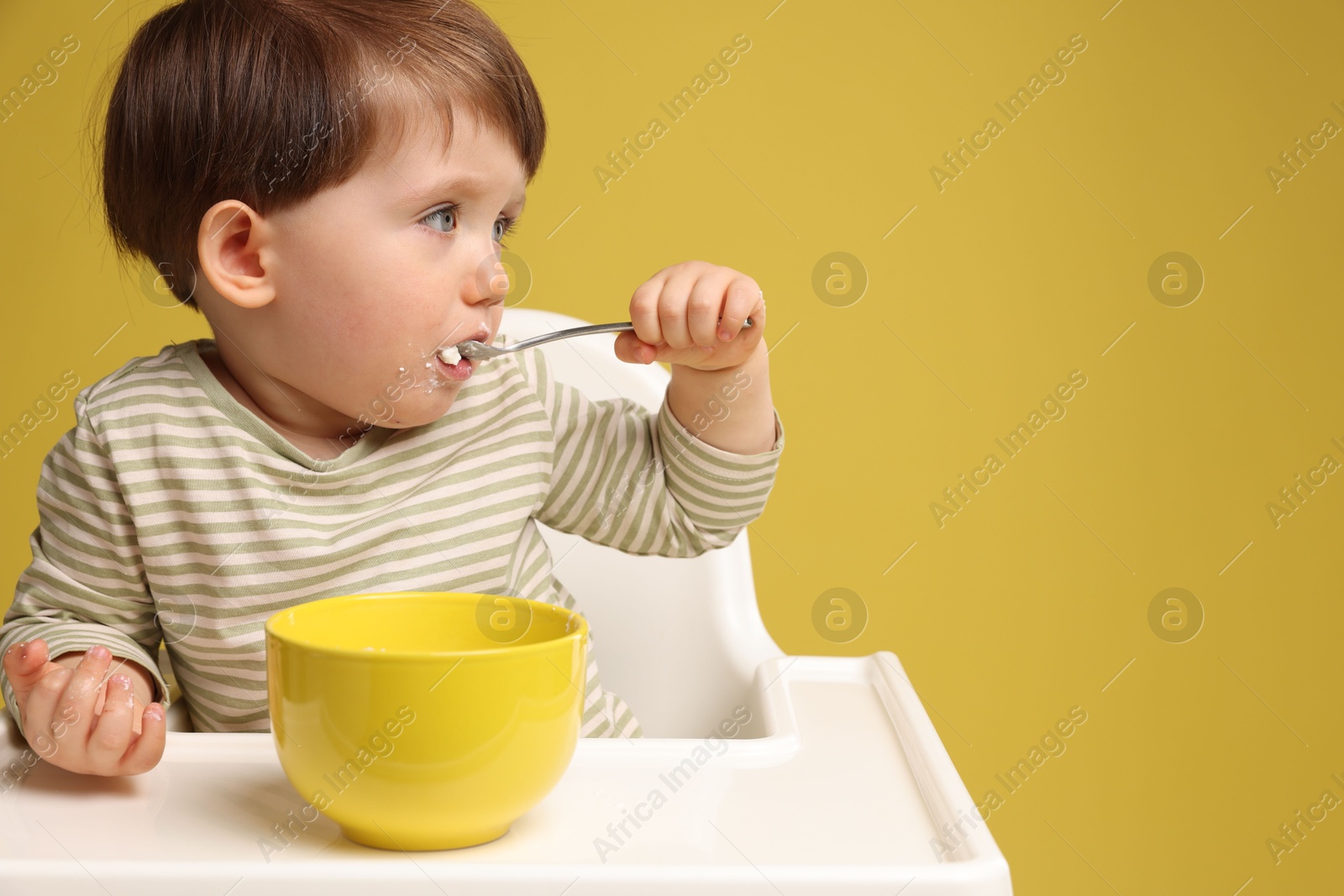 Photo of Cute little kid eating healthy baby food from bowl in high chair on yellow background, space for text