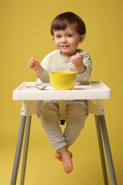 Photo of Cute little kid eating healthy baby food from bowl in high chair on yellow background