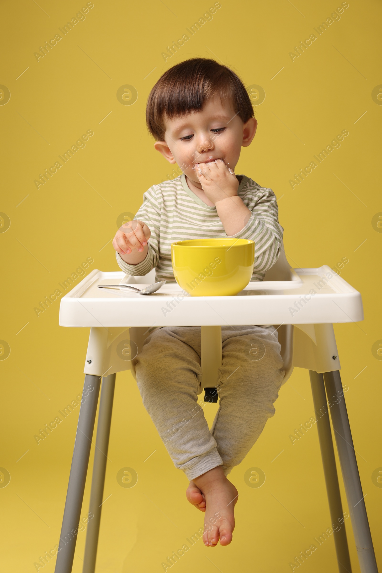 Photo of Cute little kid eating healthy baby food from bowl in high chair on yellow background