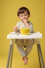 Cute little kid eating healthy baby food from bowl in high chair on yellow background