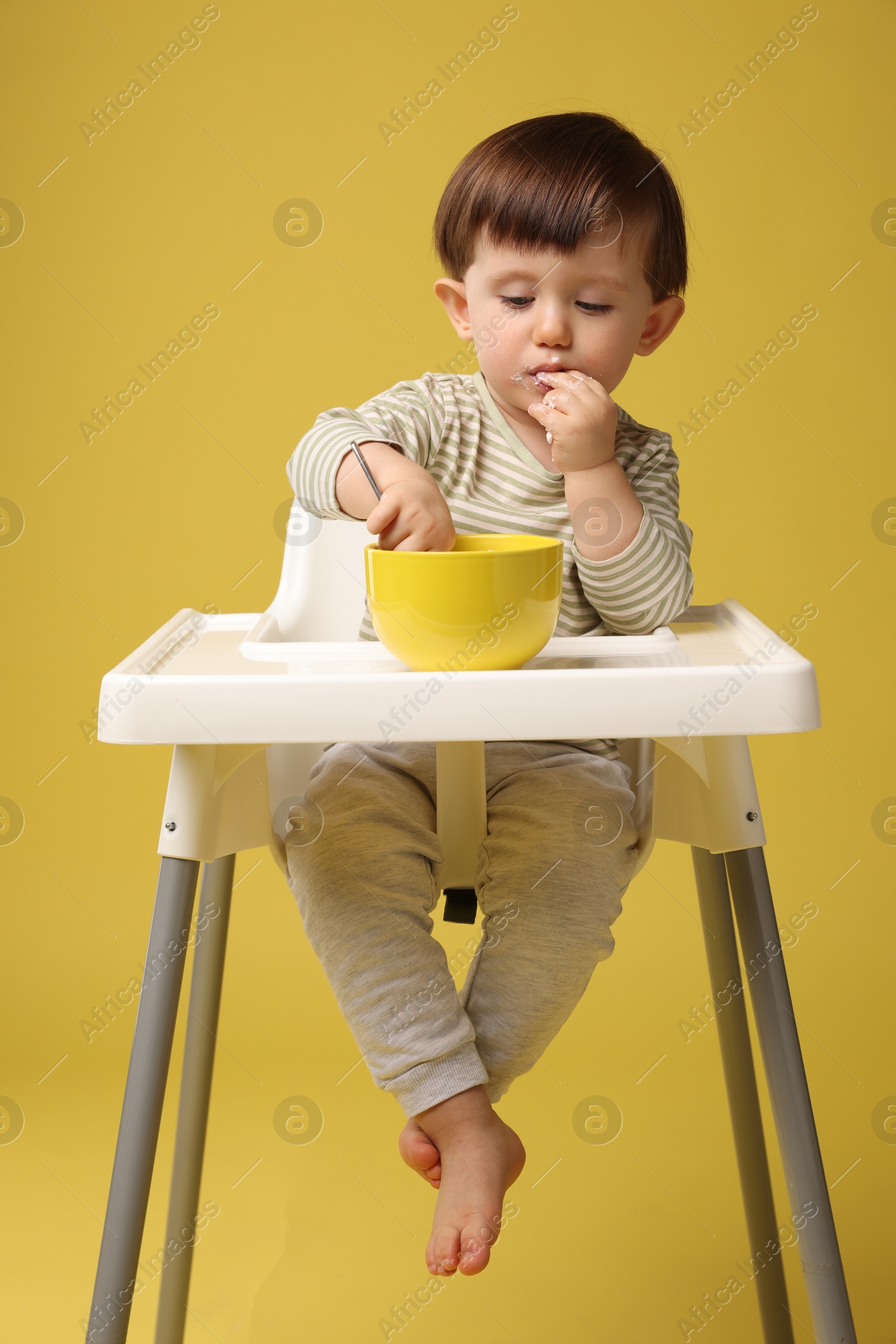 Photo of Cute little kid eating healthy baby food from bowl in high chair on yellow background