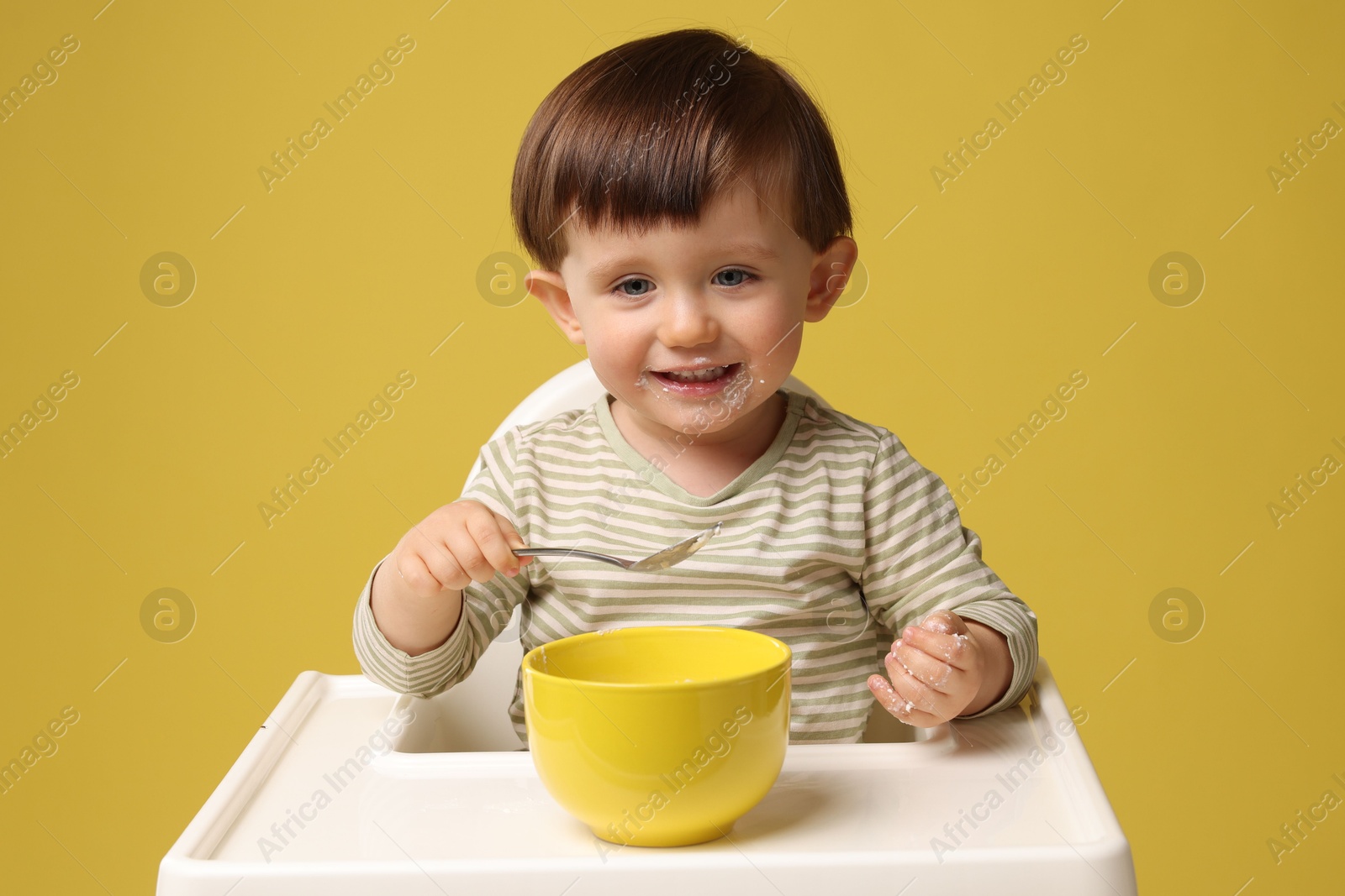 Photo of Cute little kid eating healthy baby food from bowl in high chair on yellow background