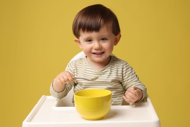 Cute little kid eating healthy baby food from bowl in high chair on yellow background