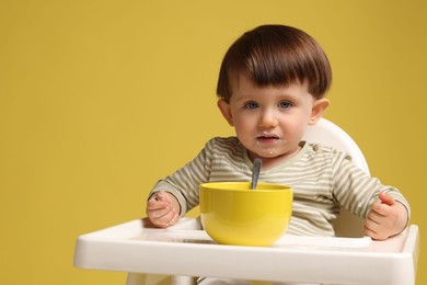 Cute little kid eating healthy baby food from bowl in high chair on yellow background