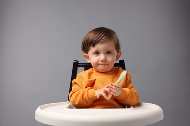 Cute little kid with spoon sitting in high chair on light grey background