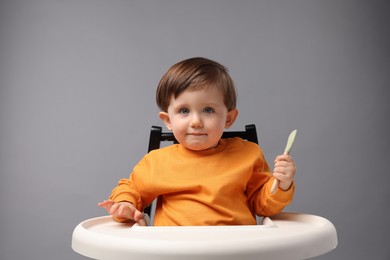 Cute little kid with spoon sitting in high chair on light grey background