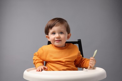 Cute little kid with spoon sitting in high chair on light grey background