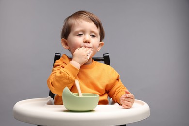 Cute little kid eating healthy baby food from bowl in high chair on light grey background