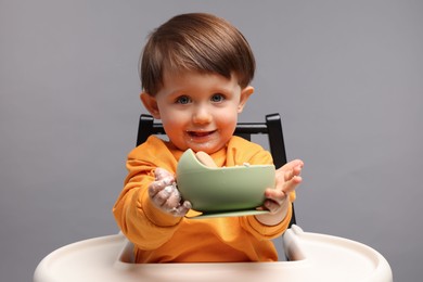 Cute little kid eating healthy baby food from bowl in high chair on light grey background